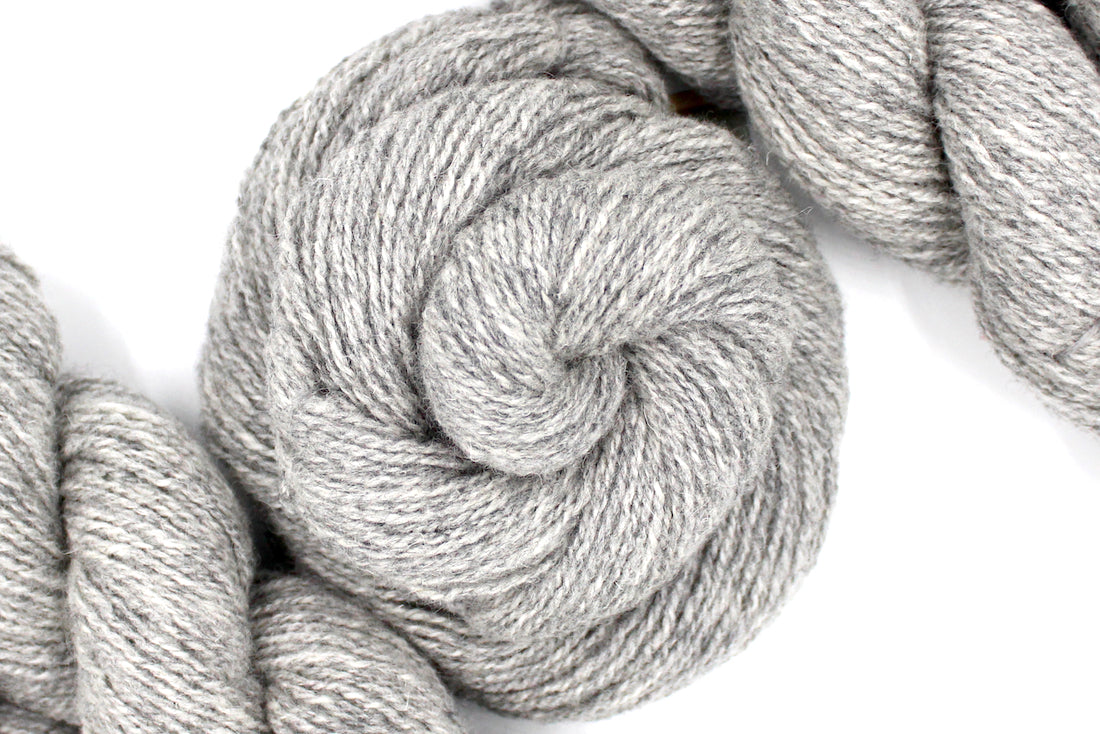 A skein of Light heathered grey, 90% Lambs wool and 10% cashmere, Dk Weight Yarn recycled by hand from unwanted sweaters swirled attractively in the center of the frame. 