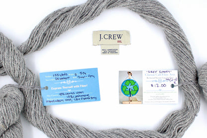 A skein of Light heathered grey, 90% Lambs wool and 10% cashmere, Dk Weight yarn encircling the sweater labels it was recycled from, as well as the hand written yarn tags detailing the specs of this recycled yarn to help you make your next project! 