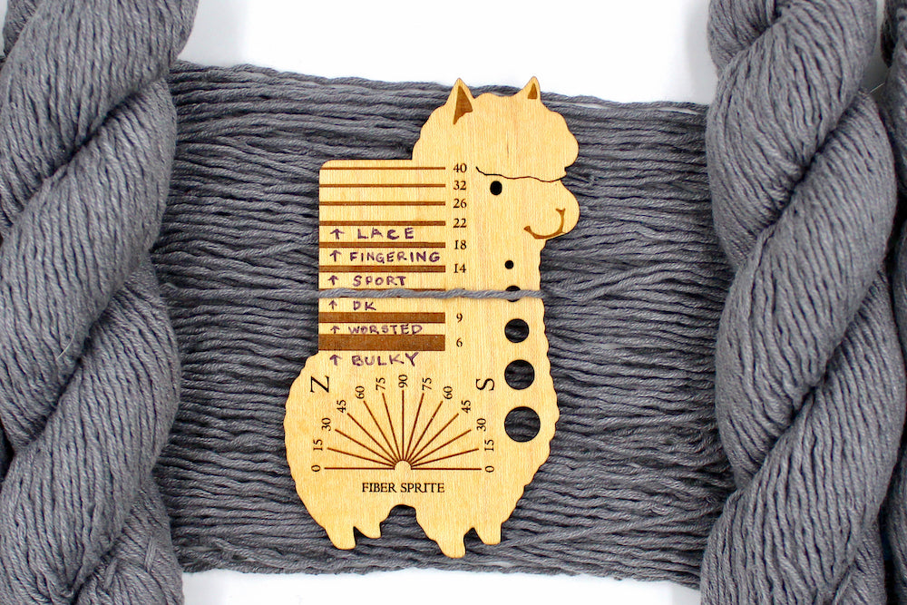 A skein of Vegan, Medium Grey, 60% Cotton, 40% Acrylic, Dk Weight Yarn recycled by hand from unwanted sweaters draped over the center of the frame with one strand on top of a Llama shaped Yarn weight tool, to better illustrate the thickness of the yarn. 