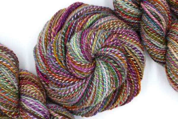 A one of a kind, hand dyed variegated skein of multicolored Pink, Orange, Gold, Blue, Green, and Purple self-striping wool Yarn coiled attractively in the center of the frame. 