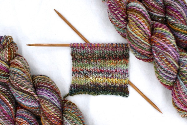 A sample swatch knitted from a one of a kind, hand dyed variegated skein of multicolored Pink, Orange, Gold, Blue, Green, and Purple self-striping wool Yarn. 