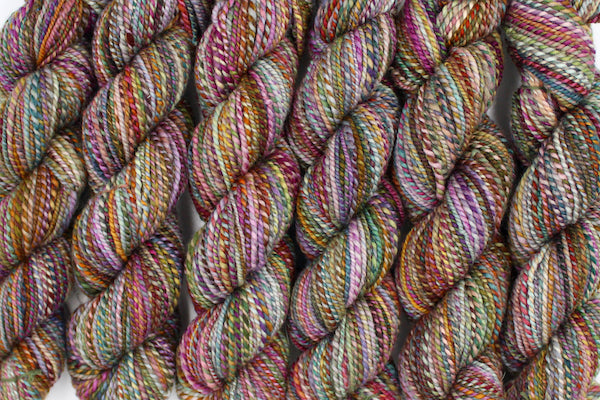 Six one of a kind, hand dyed variegated skein of multicolored Pink, Orange, Gold, Blue, Green, and Purple self-striping wool Yarn lined up side by side. 