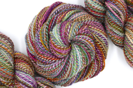 A one of a kind, Worsted Weight, hand dyed variegated skein of multicolored Pink, Orange, Gold, Blue, Green, and Purple, self-striping wool Yarn coiled attractively in the center of the frame. 