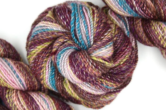 A one of a kind, hand dyed gradient skein of multicolored Brown, Maroon, Peach, Lime Green, Ballerina Pink, and Blue self-striping wool Yarn coiled attractively in the center of the frame. 