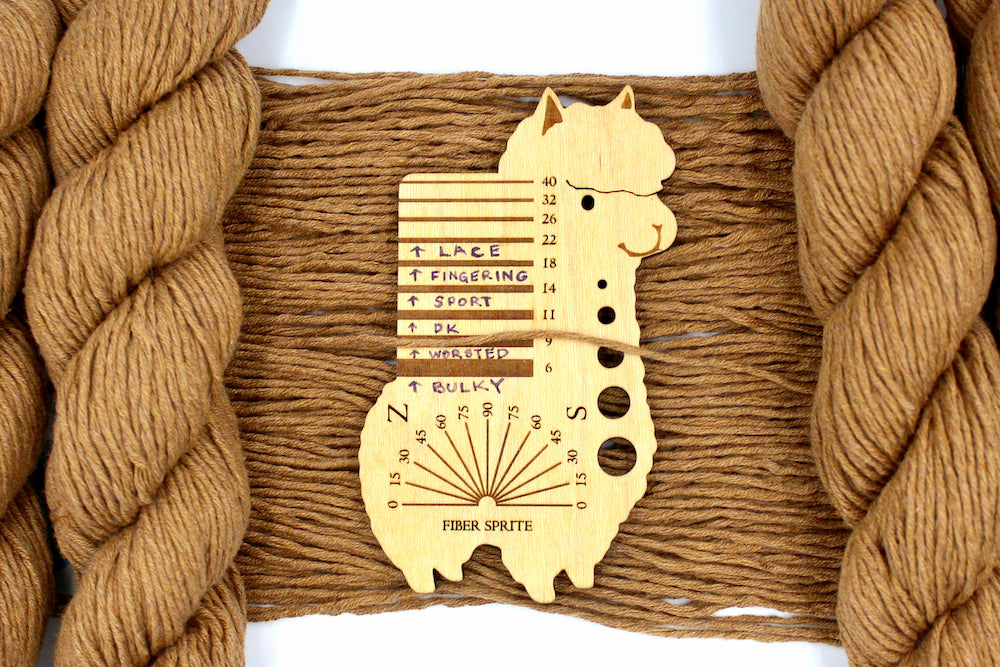 A skein of Vegan, Orangey Brown, 100% Cotton, Worsted Weight Yarn recycled by hand from unwanted sweaters draped over the center of the frame with one strand on top of a Llama shaped Yarn weight tool, to better illustrate the thickness of the yarn. 