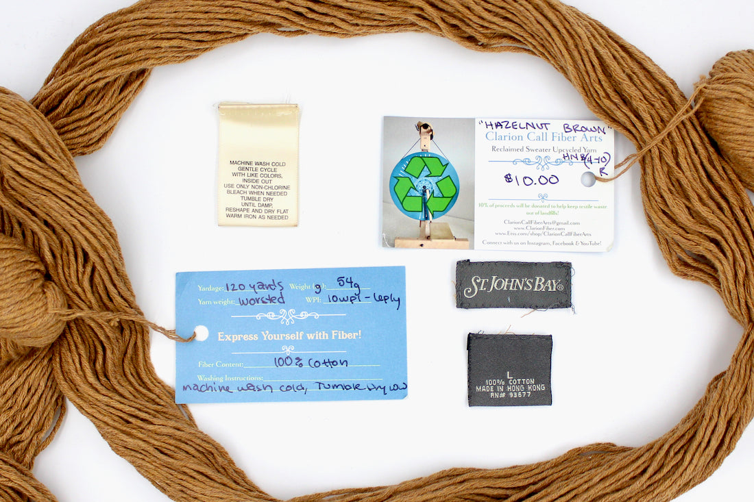 A skein of Vegan, Orangey Brown, 100% Cotton, Worsted Weight yarn encircling the sweater labels it was recycled from, as well as the hand written yarn tags detailing the specs of this recycled yarn to help you make your next project! 