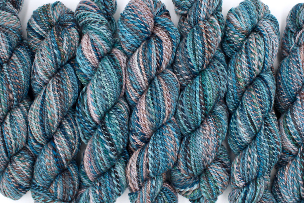 Six one of a kind, Worsted weight, hand dyed variegated skein of multicolored Blue, Teal, Taupe, and Chocolate Brown self-striping wool Yarn lined up side by side. 