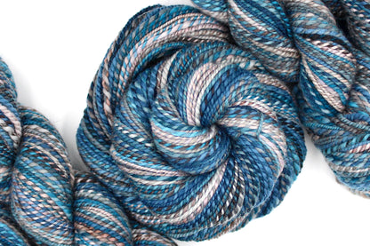 A one of a kind, Dk Weight, hand dyed variegated skein of multicolored Blue, Teal, Brown, and Taupe self-striping Hand spun Yarn, made of 100% Hand Dyed Merino Wool and coiled attractively in the center of the frame. 