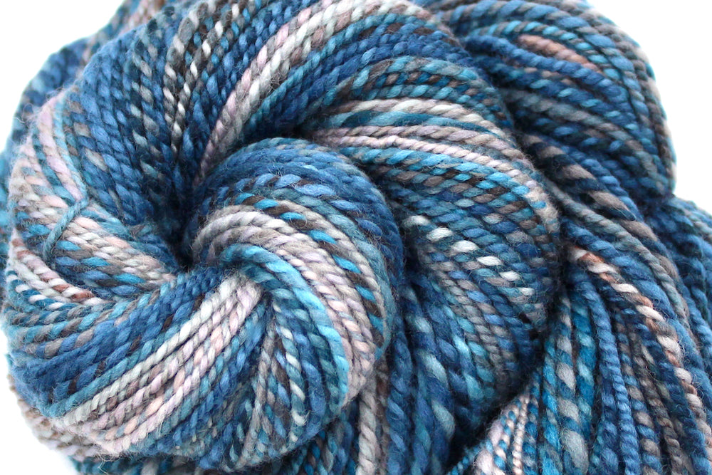 A close up view of a one of a kind, Dk Weight, hand dyed variegated skein of multicolored Blue, Teal, Brown, and Taupe self-striping Hand Spun Yarn, made of 100% Hand Dyed Merino Wool. 