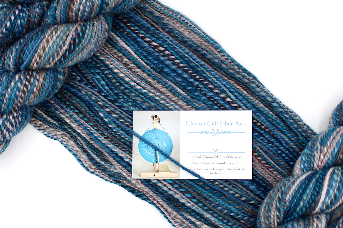 A one of a kind, Dk Weight, hand dyed variegated skein of multicolored Blue, Teal, Brown, and Taupe self-striping Hand Spun Yarn made of 100% Hand Dyed Merino Wool, draped diagonally across the frame, so you can really see the color play. 