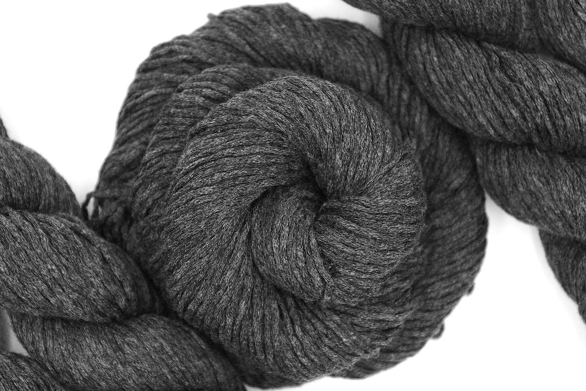 A skein of Vegan, Dark Grey, Acrylic/ Cotton, Dk weight Yarn recycled by hand from unwanted sweaters swirled attractively in the center of the frame. 