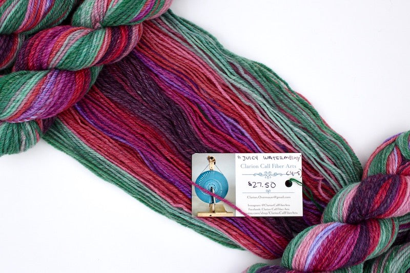 A one of a kind, hand dyed gradient skein of multicolored Plum Purple, Red, Fuchsia, Lavender, Pink, and Green self-striping wool Yarn draped diagonally across the frame, so you can really see the color play. 