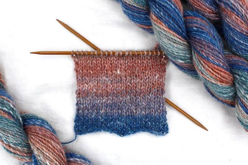 A sample swatch knitted from a one of a kind, hand dyed gradient skein of multicolored Royal Blue, Mauve, Reddish Orange, Light Teal and Dark Teal self-striping wool Yarn. 