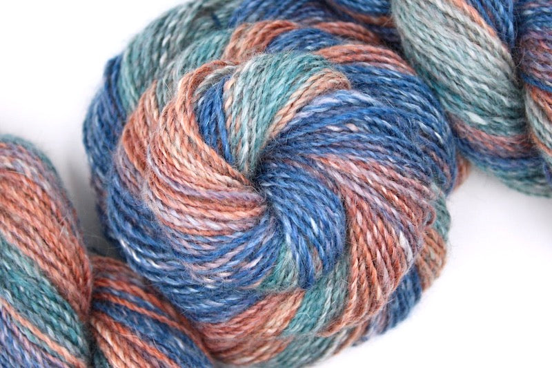 A one of a kind, hand dyed gradient skein of multicolored Royal Blue, Mauve, Reddish Orange, Light Teal and Dark Teal self-striping wool Yarn coiled attractively in the center of the frame. 