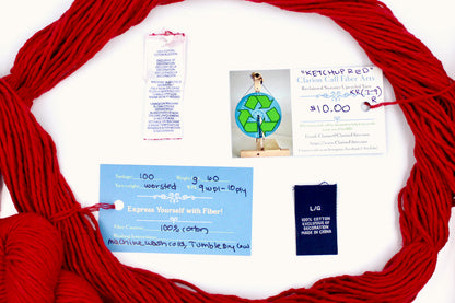 A skein of Vegan, Red, 100% Cotton, Worsted Weight yarn encircling the sweater labels it was recycled from, as well as the hand written yarn tags detailing the specs of this recycled yarn to help you make your next project! 