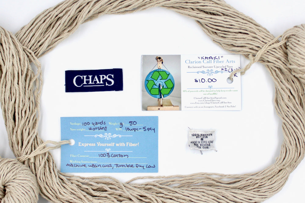 A skein of Vegan, light beige, 100% Cotton, Worsted Weight yarn encircling the sweater labels it was recycled from, as well as the hand written yarn tags detailing the specs of this recycled yarn to help you make your next project! 