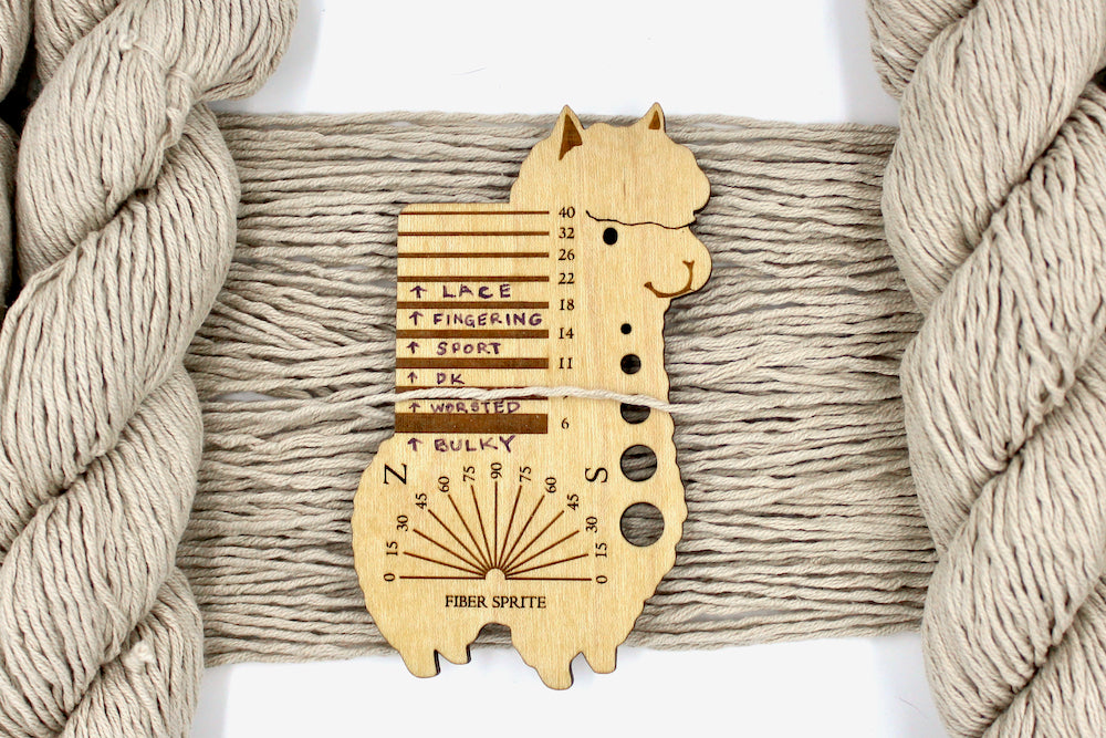 A skein of Vegan, light beige, 100% Cotton, Worsted Weight Yarn recycled by hand from unwanted sweaters draped over the center of the frame with one strand on top of a Llama shaped Yarn weight tool, to better illustrate the thickness of the yarn. 