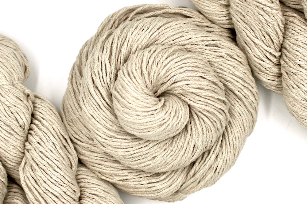 A skein of Vegan, light beige, 100% Cotton, Worsted Weight Yarn recycled by hand from unwanted sweaters swirled attractively in the center of the frame. 