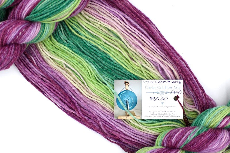 A one of a kind, hand dyed gradient skein of multicolored Magenta, Orchid Pink, Light Pink, Light Green, Lime Green, and Kelly Green self-striping wool Yarn draped diagonally across the frame, so you can really see the color play. 