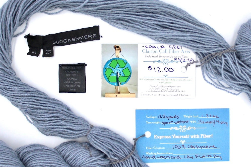A skein of Light Silver Grey, 100% Cashmere wool, Sport Weight yarn encircling the sweater labels it was recycled from, as well as the hand written yarn tags detailing the specs of this upcycled yarn to help you make your next project! 