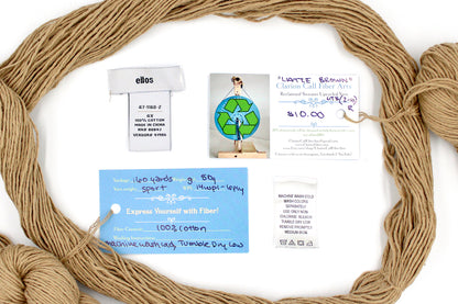 A skein of Vegan, golden tan, 100% Cotton, Sport Weight yarn encircling the sweater labels it was recycled from, as well as the hand written yarn tags detailing the specs of this recycled yarn to help you make your next project! 