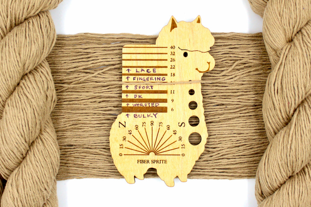 A skein of Vegan, golden tan, 100% Cotton, Sport Weight Yarn recycled by hand from unwanted sweaters draped over the center of the frame with one strand on top of a Llama shaped Yarn weight tool, to better illustrate the thickness of the yarn. 