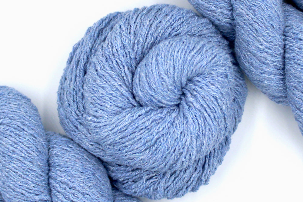 A skein of Vegan, baby blue, upcycled Cotton, Polyester, and acrylic, Sport Weight Yarn recycled by hand from unwanted sweaters swirled attractively in the center of the frame. 