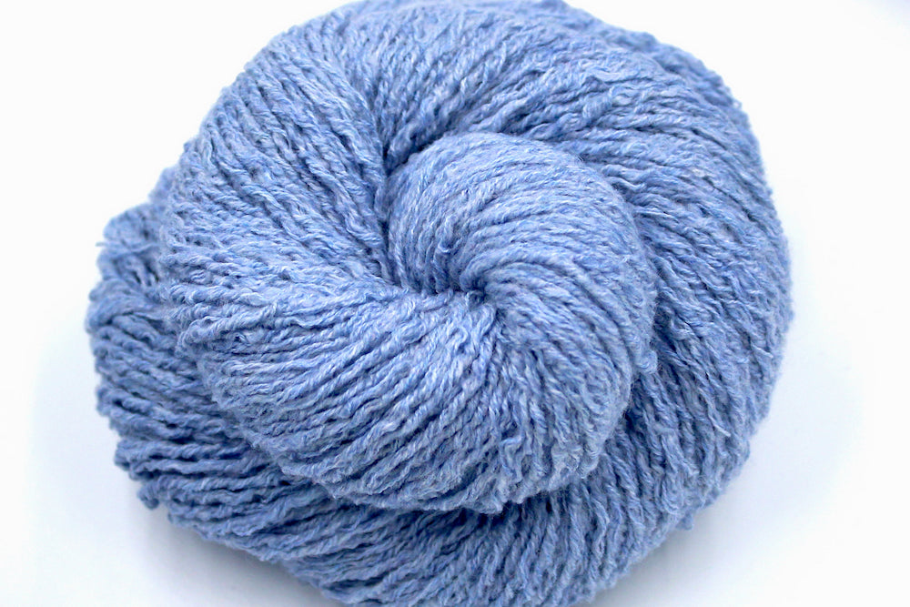 A close up shot of a skein of Vegan, baby blue, upcycled Cotton, Polyester, and acrylic, Sport Weight Yarn recycled by hand from unwanted sweaters beautifully coiled in the center of the frame. 