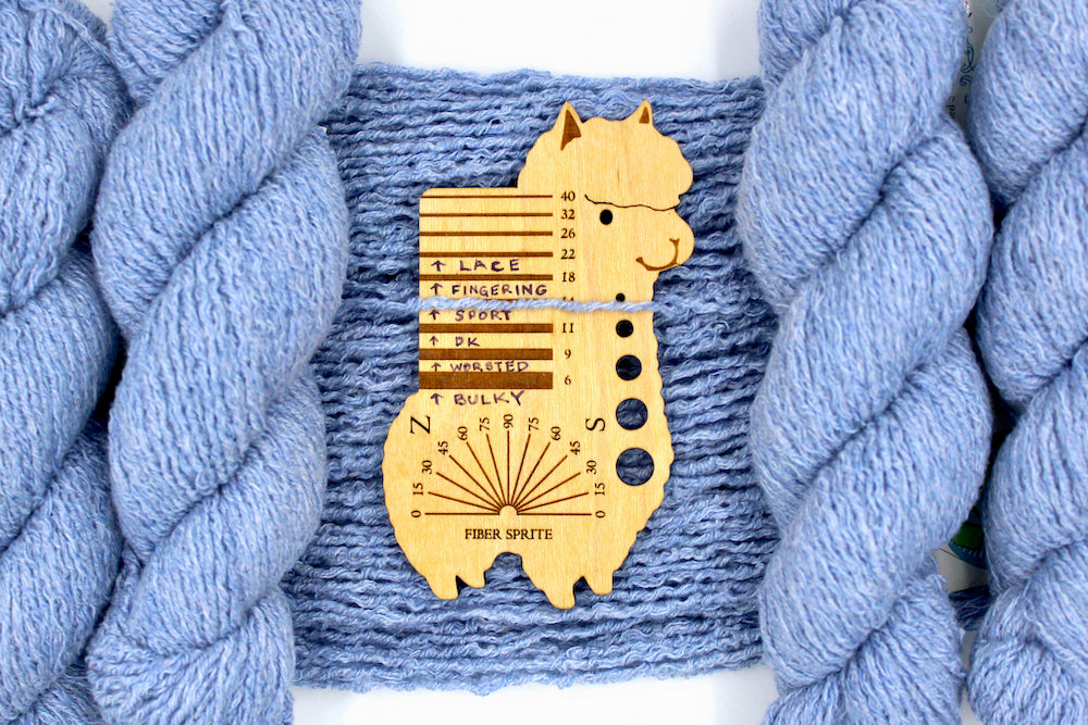 A skein of Vegan, baby blue, upcycled Cotton, Polyester, and acrylic, Sport Weight Yarn recycled by hand from unwanted sweaters draped over the center of the frame with one strand on top of a Llama shaped Yarn weight tool, to better illustrate the thickness of the yarn. 