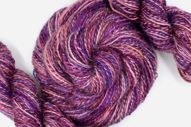 A one of a kind, hand dyed variegated skein of multicolored Peach, Pink, Purple, and Magenta self-striping wool Yarn coiled attractively in the center of the frame. 