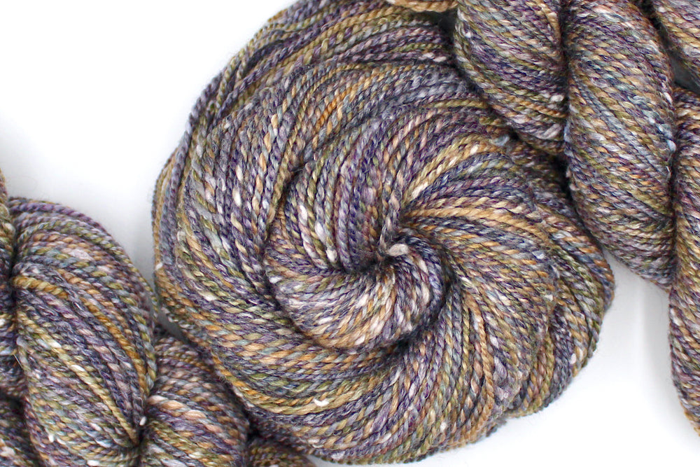 A one of a kind Dk Weight, hand dyed variegated skein of multicolored Grey, Purple, Olive Green, and Gold self-striping Hand spun Yarn, made with Tweed, Wool and Bamboo; coiled attractively in the center of the frame. 
