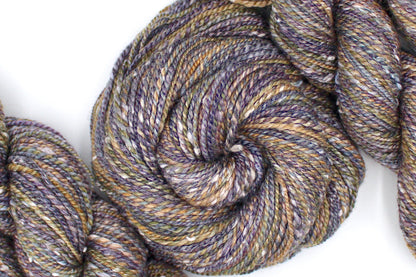 A one of a kind Dk Weight, hand dyed variegated skein of multicolored Grey, Purple, Olive Green, and Gold self-striping Hand spun Yarn, made with Tweed, Wool and Bamboo; coiled attractively in the center of the frame. 