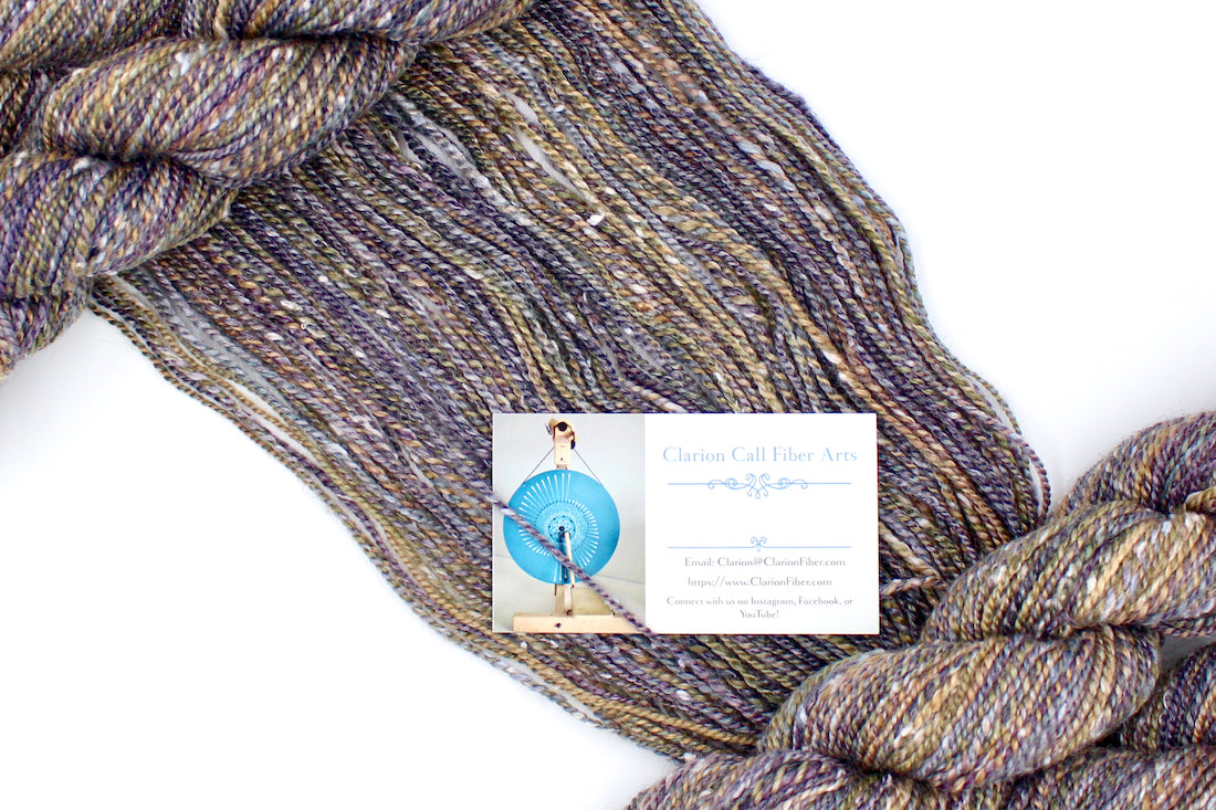 A one of a kind, Dk Weight, hand dyed variegated skein of multicolored Grey, Purple, Olive Green, and Gold self-striping Hand spun Yarn, made with Tweed, Wool and Bamboo, draped diagonally across the frame, so you can really see the color play. 