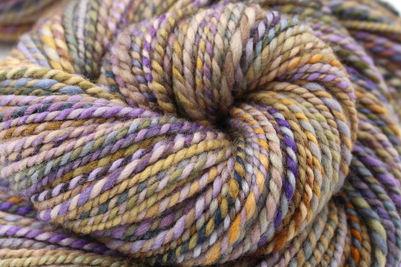 A close up view of a one of a kind, variegated skein of multicolored Purple, Lavender, Periwinkle, Taupe, Gold, and Orange self-striping wool Yarn. 
