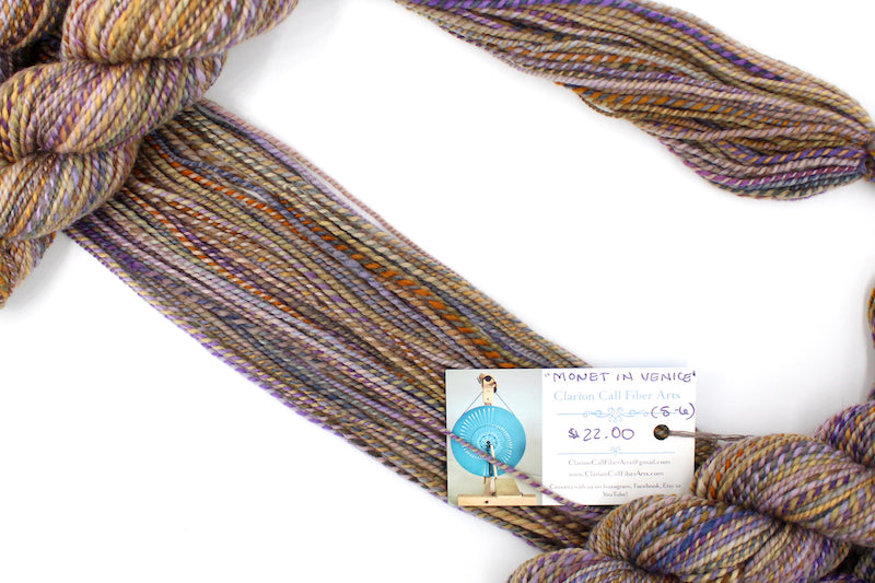 A one of a kind, hand dyed variegated skein of multicolored Purple, Lavender, Periwinkle, Taupe, Gold, and Orange self-striping wool Yarn draped diagonally across the frame, so you can really see the color play. 