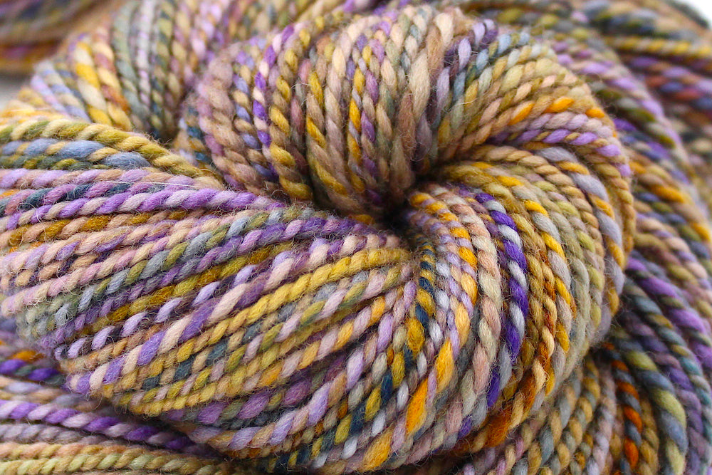 A close up view of a one of a kind, Dk Weight, hand dyed Variegated skein of multicolored Purple, Lavender, Periwinkle, Taupe, Gold, and Orange self-striping Yarn, made of 100% Hand Dyed Merino Wool. 