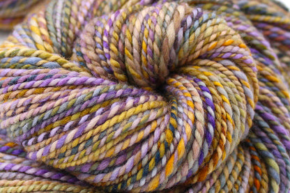 A close up view of a one of a kind, Dk Weight, hand dyed Variegated skein of multicolored Purple, Lavender, Periwinkle, Taupe, Gold, and Orange self-striping Yarn, made of 100% Hand Dyed Merino Wool. 