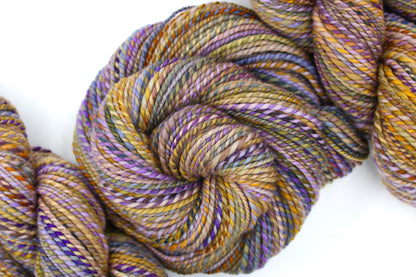 A one of a kind, Dk Weight, hand dyed variegated skein of multicolored Purple, Lavender, Periwinkle, Taupe, Gold, and Orange self-striping Yarn, made of 100% Hand Dyed Merino Wool and coiled attractively in the center of the frame. 