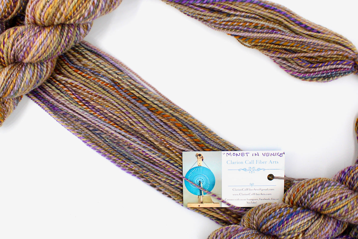 A one of a kind, Dk Weight, hand dyed Variegated skein of multicolored Purple, Lavender, Periwinkle, Taupe, Gold, and Orange self-striping Yarn made of 100% Hand Dyed Merino Wool, draped diagonally across the frame, so you can really see the color play. 
