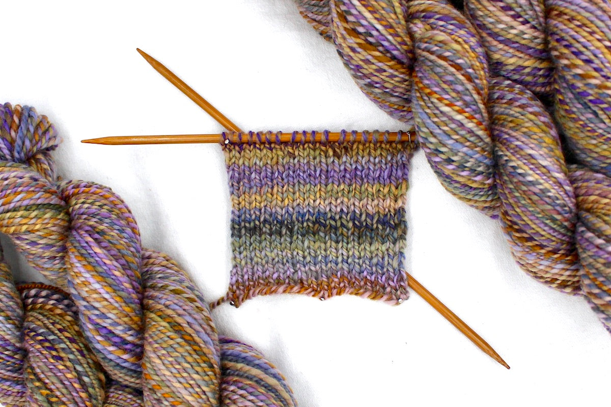 A sample swatch knitted on size 5 knitting needles from a one of a kind, Dk Weight, hand dyed Variegated skein of self striping Maroon, Pink, Cornflower Blue, and Lime Gr multicolored Purple, Lavender, Periwinkle, Taupe, Gold, and Orange self-striping Yarn, made of 100% Hand Dyed Merino Wool. 