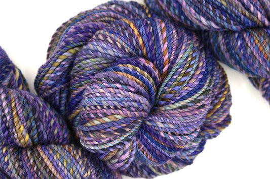 A one of a kind, hand dyed variegated skein of multicolored Purple, Blue, Green, Pink and Gold self-striping wool Yarn coiled attractively in the center of the frame. 