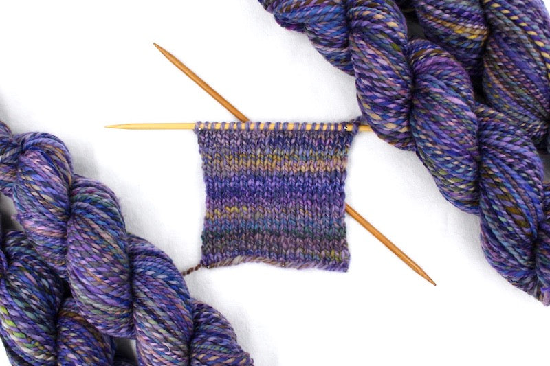A sample swatch knitted from a one of a kind, hand dyed variegated skein of multicolored Purple, Blue, Green, Pink and Gold self-striping wool Yarn. 