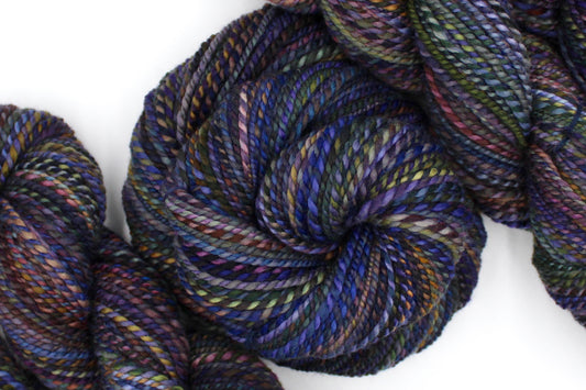 A one of a kind, Worsted Weight, hand dyed variegated skein of multicolored Blue, Purple, Yellow, and Olive Green, and Maroon, self-striping wool Yarn coiled attractively in the center of the frame. 