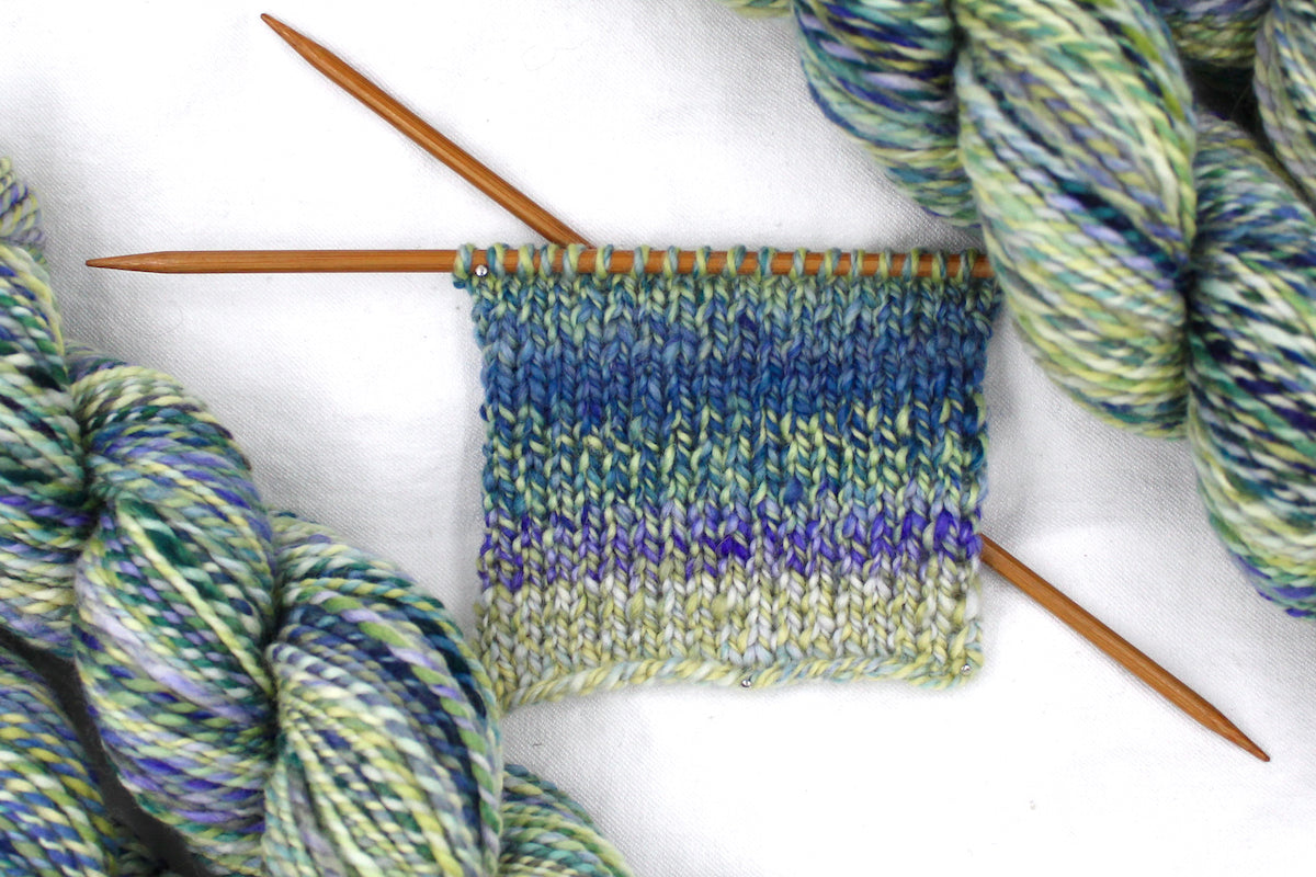 A sample swatch knitted from a one of a kind, Worsted weight, hand dyed variegated skein of multicolored Blue, Green, Teal, and Light Yellow self-striping wool Yarn. 