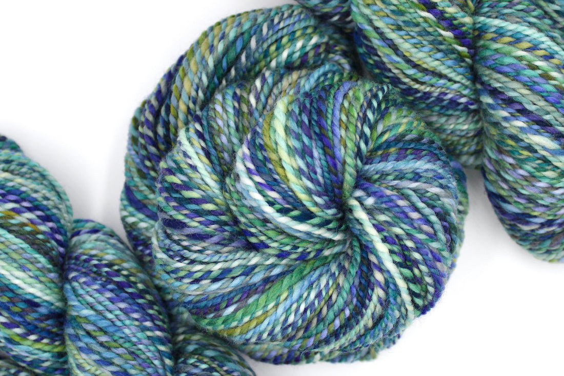 A one of a kind, Worsted Weight, hand dyed variegated skein of multicolored Blue, Green, Teal, Light Yellow, and Lime Green, self-striping wool Yarn coiled attractively in the center of the frame. 