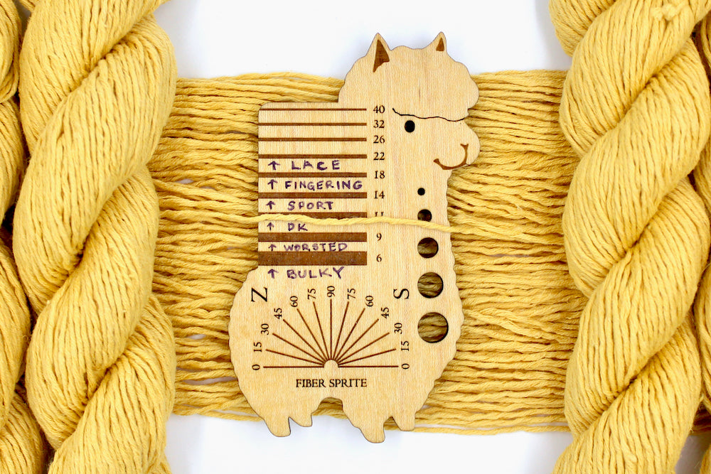 A skein of Vegan, Mustard Yellow, Cotten blend, DK Weight Yarn recycled by hand from unwanted sweaters draped over the center of the frame with one strand on top of a Llama shaped Yarn weight tool, to better illustrate the thickness of the yarn. 