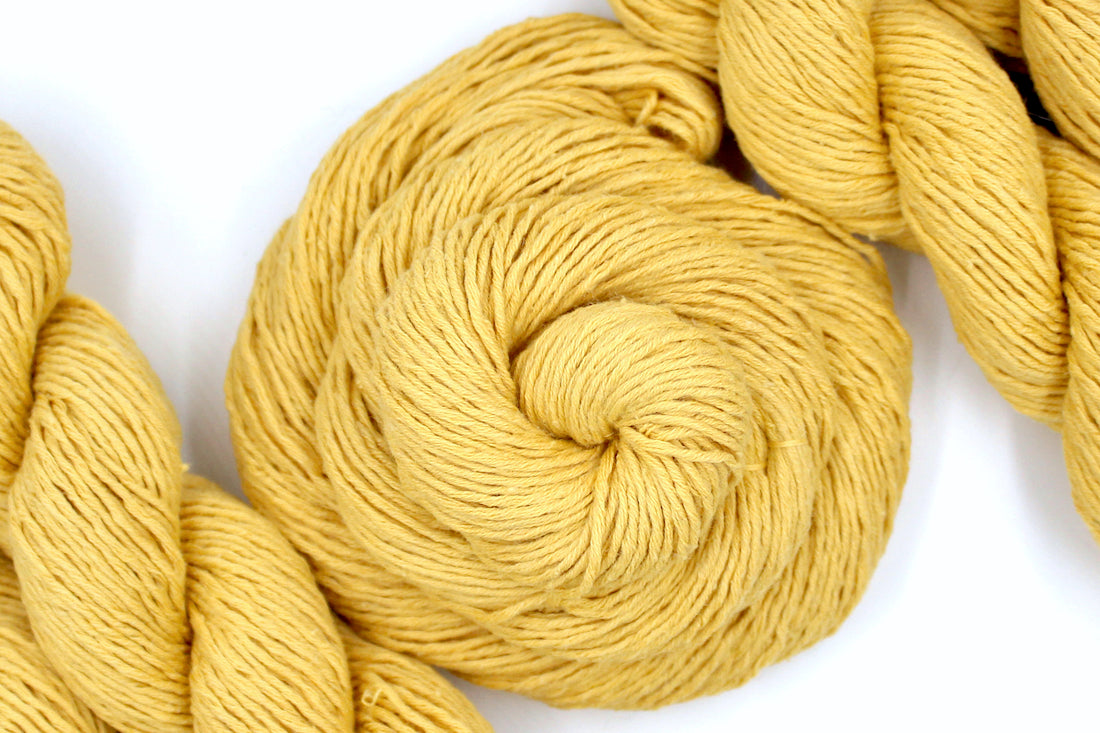 A skein of Vegan, Mustard Yellow, Cotten blend, DK Weight Yarn recycled by hand from unwanted sweaters swirled attractively in the center of the frame. 