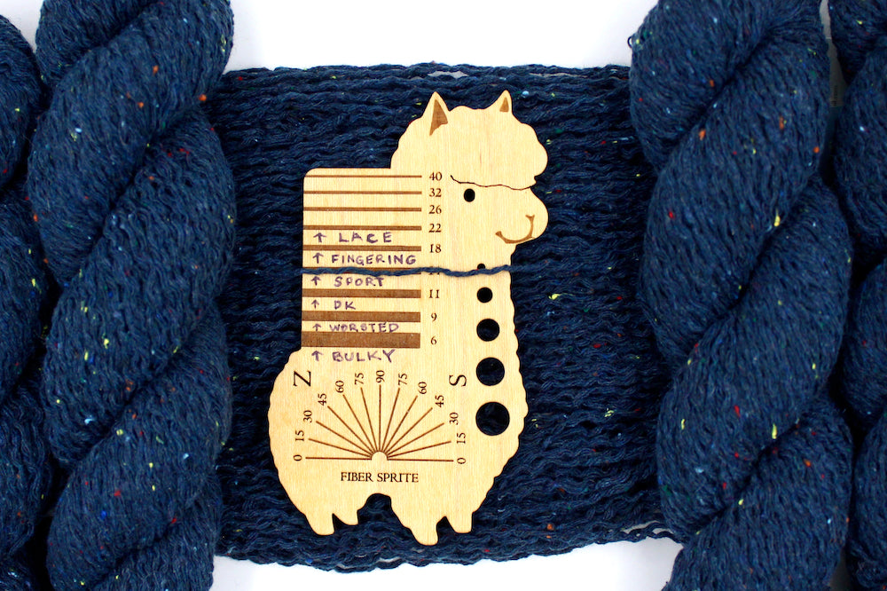 A skein of Dark Blue and Rainbow Tweed, Wool and Acrylic, Sport Weight Yarn recycled by hand from unwanted sweaters draped over the center of the frame with one strand on top of a Llama shaped Yarn weight tool, to better illustrate the thickness of the yarn. 