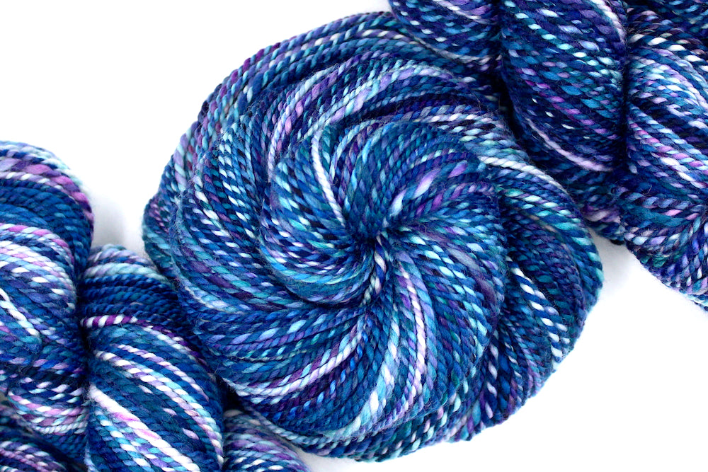 A one of a kind, Dk Weight, hand dyed variegated skein of multicolored Blue, and Purple self-striping Hand spun Yarn, made of 100% Hand Dyed Merino Wool and coiled attractively in the center of the frame. 
