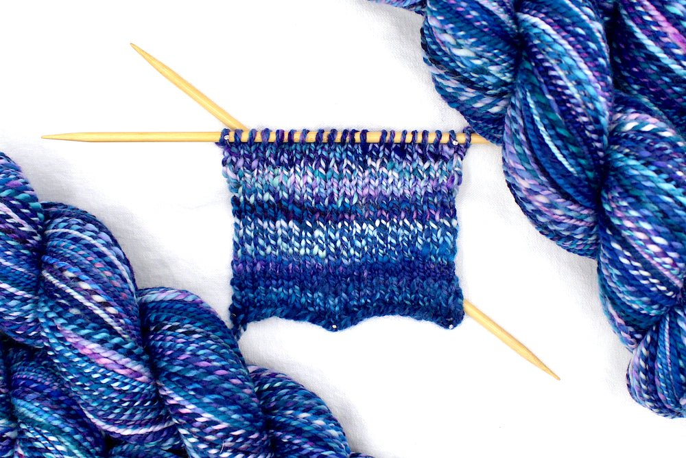 A sample swatch knitted on size 5 knitting needles from a one of a kind, Dk Weight, hand dyed variegated skein of multicolored Blue, and Purple self-striping Hand spun Yarn, made of 100% Hand Dyed Merino Wool. 
